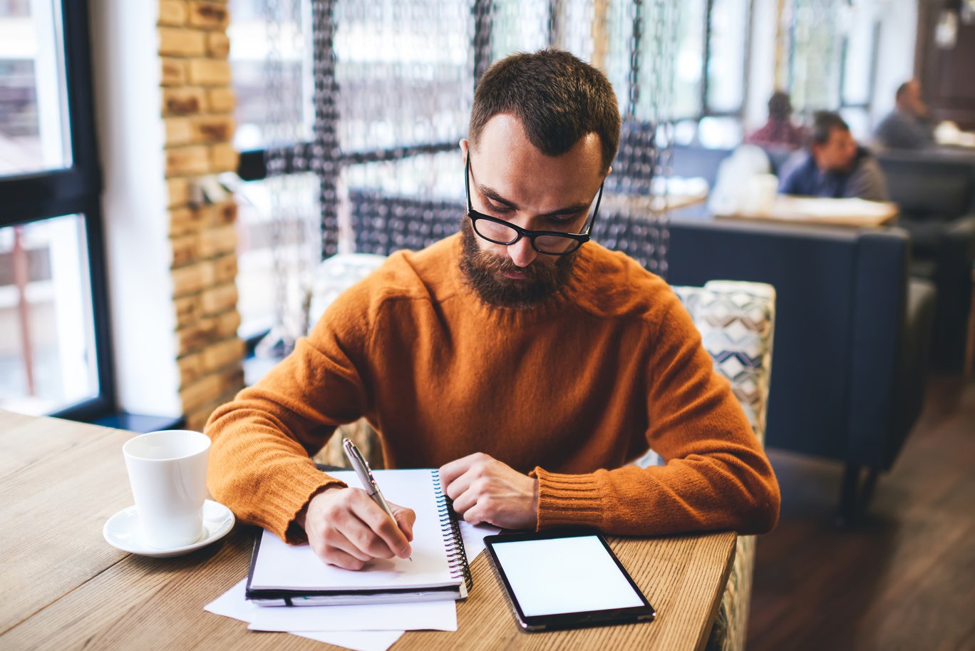 Bearded male writer writing in notebook in cafe at daytime